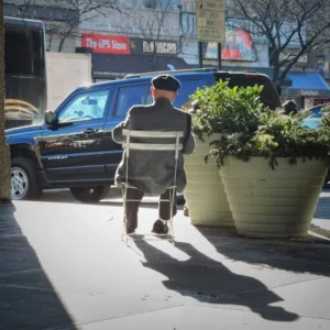 Elderly man sitting on a chair on a sidewalk in Queens, NY